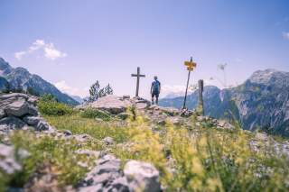 Mann steht an Gipfelkreuz und genießt die Aussicht