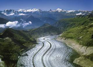 Areal view über eine sich durch die Landschaft windende Gletscherzunge, im Hintergrund hohe Berggipfel.