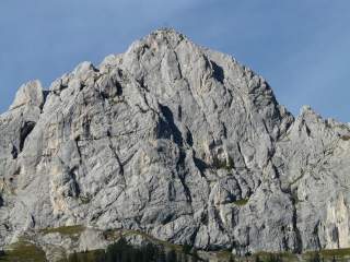 Das Bild gibt einen freien Blick auf die Südwand der Roten Flüh, ein Berg der Tannheimer Berge im Allgäu. Der Fels ist frei von Vegetation und der Himmel im Hintergrund blau. Auf dem Gipfel des Berges befindet sich ein Gipfelkreuz.