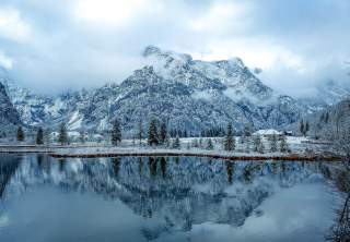 Verschneite Berge mit Tannen und Häusern am Ufer eines ruhigen Sees, in dem sich die Landschaft spiegelt.