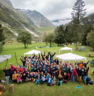 Eine große Gruppe von Menschen steht und sitzt fröhlich auf einer Wiese mit Zelten und Pavillons, umgeben von Bergen und Bäumen. Alle wirken jubelnd und genießen die Natur.