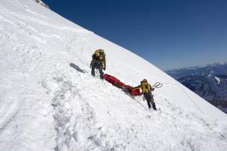 Zwei Bergsteiger in gelber Ausrüstung transportieren eine rot verpackte Person auf einer Trage einen verschneiten Hang hinauf, im Hintergrund schneebedeckte Berge.