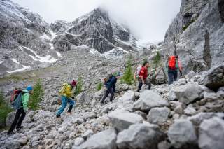 Fünf Wanderer mit Rucksäcken steigen einen steinigen Hang in einer felsigen, schneebedeckten Berglandschaft hinauf.