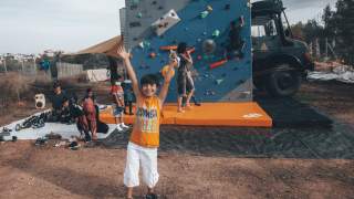 Ein Junge streckt freudig die Hände Richtung Himmel, im Hintergrund steht die mobile Boulderwand. Einige Kinder stehen drum herum oder hängen an der Wand.