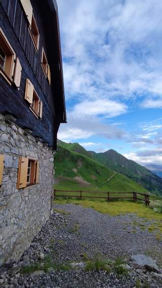Stein- und Holzhaus mit geöffneten Fensterläden am Rande eines grünen Bergpanoramas unter blauem Himmel mit Wolken.