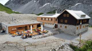 Bergsteigerhütte mit mehreren Holztischen und Sitzbänken im Freien, umgeben von felsigem Gebirge. Menschen sitzen an den Tischen und genießen die Berglandschaft.