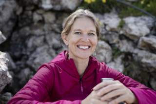 Nahaufnahme einer lächelnden Frau in beerenfarbener Outdoorjacke vor Felsen, die eine Aluflasche hält.