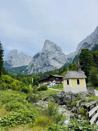 Kleine gelbe Kapelle auf einem Felsen neben einem Bach mit grüner Vegetation, dahinter ein traditionelle Berghütte und steile, felsige Berge unter bewölktem Himmel.