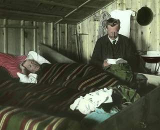 Zwei Männer im Matratzenlager der Amberger Hütte um 1910. Einer liegt im Bett, der andere sitzt daneben und blickt in seine Brotdose.
