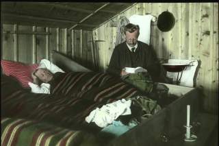 Zwei Männer um 1910 im Matratzenlager der Amberger Hütte. Einer liegt zugedeckt im Bett, der andere sitzt daneben und blickt in seine Brotdose.