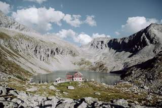 Steinhütte mit roten Fensterläden an einem Bergsee, umgeben von felsigen, grauen Bergen unter blauem Himmel mit Wolken.