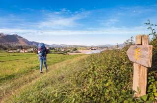 Eine wandernde Person mit Rucksack geht auf einem Pfad durch eine grüne Landschaft mit Bergen und einem Dorf im Hintergrund, rechts ein hölzerner Wegweiser mit gelbem Pilgerzeichen.