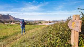Eine wandernde Person mit Rucksack geht auf einem Pfad durch eine grüne Landschaft mit Bergen und einem Dorf im Hintergrund, rechts ein hölzerner Wegweiser mit gelbem Pilgerzeichen.