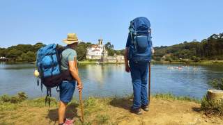 Zwei Wanderer schauen über einen breiten Fluss hinüber zu einer weiß leuchtenden Kirche.