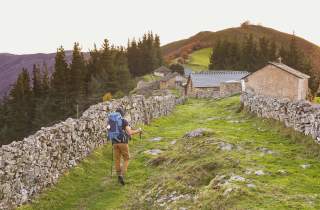 Ein Wanderer mit großem Rucksack und Wanderstöcken geht auf einem von niedrigen Steinmauern gesäumten Pfad zu einer kleinen Ansiedlung aus Steinhäusern in einer grünen, hügeligen Landschaft.
