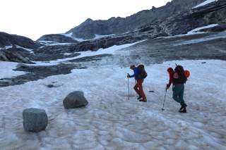 Zwei Bergsteiger laufen über einen Gletscher mit Firnauflage und Felsbrocken.