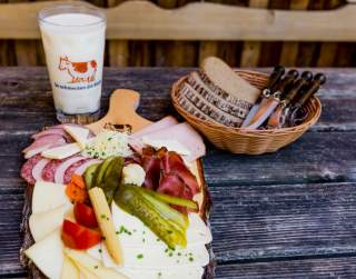 Brotzeitbrett mit verschiedenen Käsesorten, Wurst und eingelegtem Gemüse, dazu ein Glas Milch und ein Brotkorb mit mehreren Messern auf einem rustikalen Holztisch.