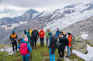 Eine Gruppe von Wanderern steht auf einem alpinen Pfad vor schneebedeckten Bergen und Gletschern, einige tragen Rucksäcke und Wanderstöcke. Der Himmel ist bewölkt und die Szenerie wirkt kühl. Ein man in der Gruppe hebt die Hand und scheint den anderen teilnehmenden etwas zu erklären.