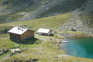 Berghütte mit Nebengebäuden an einem kleinen See, umgeben von Wiese, Felsen und Bergen; einige Menschen sitzen am Ufer.