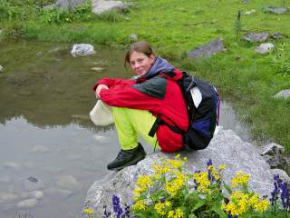 Eine Person in Outdoor-Kleidung sitzt auf einem Felsen am Wasser, mit Rucksack, umgeben von grüner Wiese und gelben sowie violetten Blumen.
