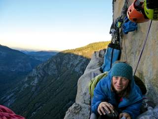 Frau in Kletterausrüstung liegt auf einem schmalen Felsvorsprung an einer steilen Felswand, mit Ausrüstung und Helmen im Hintergrund, Blick auf bewaldete Berge im Sonnenlicht.
