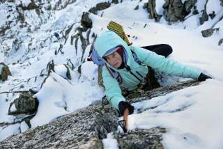 Eine Person klettert mit Eispickel und Winterausrüstung eine verschneite Felswand hinauf.