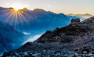 Eine Hütte mitten im Hochgebirge, hinter dem Bergkamm kommt die Sonne hervor.