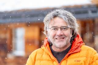 Porträt eines lächelnden Mannes mit Brille und grauen, lockigen Haaren, der draußen bei leichtem Schneefall eine orange Winterjacke trägt.