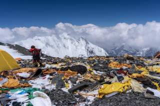 Bergsteiger steht auf einem schneebedeckten Berghang, umgeben von Müll, Zelten und Ausrüstung; im Hintergrund schneebedeckte Gipfel und Wolken.