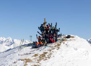 Eine Gruppe junger Skitourengänger*innen steht am Gipfelkreuz und jubelt in die Kamera