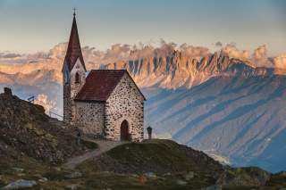 Kleine, steinerne Kirche mit spitzem Turm steht auf einem Berghang, im Hintergrund schroffe, sonnenbeschienene Berge und Wolken.