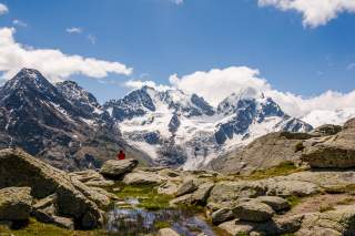 Blick auf die vergletscherte Berninagruppe, im Vordergrund sitz eine Person in roter Jacke auf einem Fels, den Rücken zur Kamera gewandt.