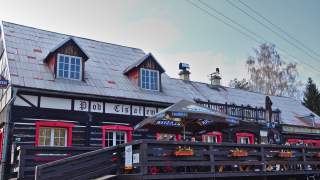 Ein großes, altes Haus mit Fachwerk im Obergeschoss und dunklem Holz. Davor auf der Terrasse sitzen Menschen beim Essen.