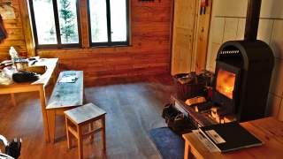 Im inneren einer Hütte, rechts brennt der Kamin, links sieht man einen großen Tisch und Holzbänke.