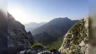 Mann klettert vorsichtig an steiler Felswand in gebirgiger Landschaft mit Rucksack und orangefarbener Stirnband.
