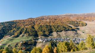 Blick auf das Dorf am Talhang, rundherum sieht man gelblich gefärbten Wald und trockene Wiesen.