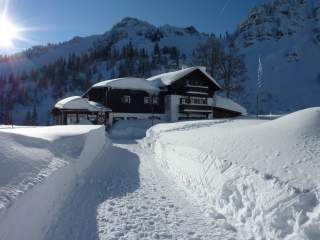Verschneites Hütte in den Bergen mit einem freigeräumten Weg, umgeben von hohen Schneehügeln und sonnigem Himmel.