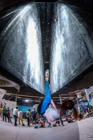 Eine Person klettert kopfüber an einer Überhang-Kletterwand in einer Kletterhalle, während mehrere Leute im Hintergrund zuschauen.