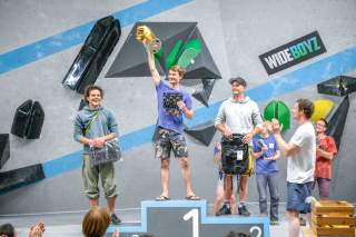 Drei Männer stehen auf einem Siegerpodest bei einem Kletterwettbewerb, der Erstplatzierte hält einen Pokal in die Luft, während Menschen im Hintergrund applaudieren.