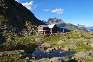 Eine Hütte aus Holz vor hochalpinen Bergen, davor ein kleiner See in dem sich die Hütte spiegelt.