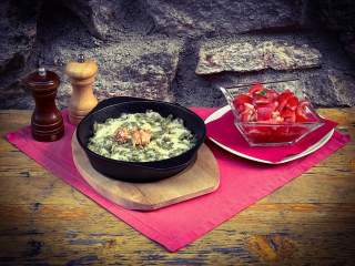 Brennnesselspätzle mit Käse und Röstzwiebeln in einer schwarzen Pfanne, daneben ein Salat, Salz- und Pfeffermühle