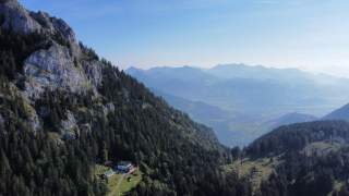 Blick auf bewaldete Berghänge mit einer Hütte einer weitläufigen Berglandschaft im Hintergrund