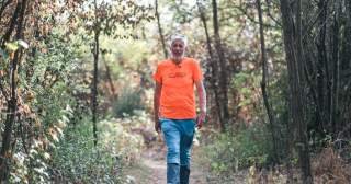 Älterer Mann mit grauen Haaren und Bart geht auf einem Waldweg, umgeben von Bäumen und herbstlicher Vegetation. Er trägt ein sportliches Outfit: oranges T-Shirt und blaue Berghose.