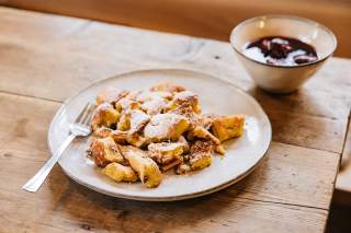 Auf einem Holztisch steht ein Teller mit Kaiserschmarrn, daneben eine Schussel mit Rotweinzwetschgen