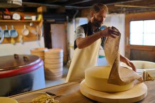 Ein Mann in Schürze bearbeitet in einer Käserei einen großen runden Laib Käse und hebt ein Tuch davon ab.