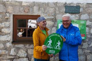 Zwei Männer stehen lachend vor einer Steinwand und halten ein rundes grünes Schild mit der Aufschrift 