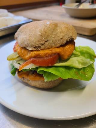 Burgerbrötchen mit Salat, Tomate, Zwiebel, Gewürzgurke und einer orangefarbenen Sauce auf einem weißen Teller.