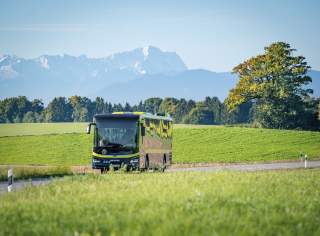 Ein Reisebus fährt auf einer Landstraße durch grüne Wiesen, im Hintergrund sind Berge und ein großer Baum zu sehen.