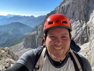 Mann mit rotem Kletterhelm und Rucksack macht ein Selfie in felsiger Berglandschaft mit blauem Himmel und entfernten Bergen im Hintergrund.