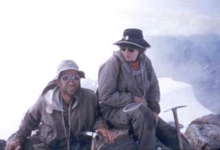 Zwei Personen in Bergsteigerausrüstung sitzen auf felsigem Gelände mit Berglandschaft im Hintergrund, jeweils mit Sonnenbrille und Hut, beide mit Felspickel.